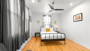 A bedroom with a bed, a ceiling fan, a bedside table, a nightstand, and a window with curtains.