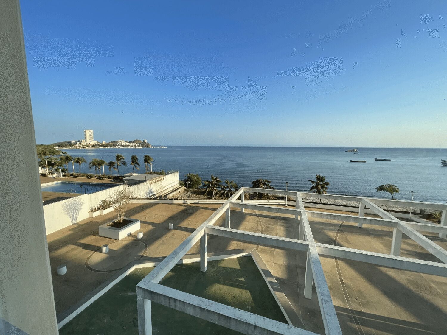 A view from a balcony overlooking a pool area with a clear view of the sea and distant buildings.
