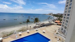 A view of a beach with a pool and lounge chairs, boats in the water, and a building in the background. - Seafront Living, Great for families and friends traveling together (Pampatar)