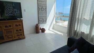 Una sala de estar con televisor, un tapiz decorativo, un aparador de madera y vistas al mar a través de amplias ventanas.