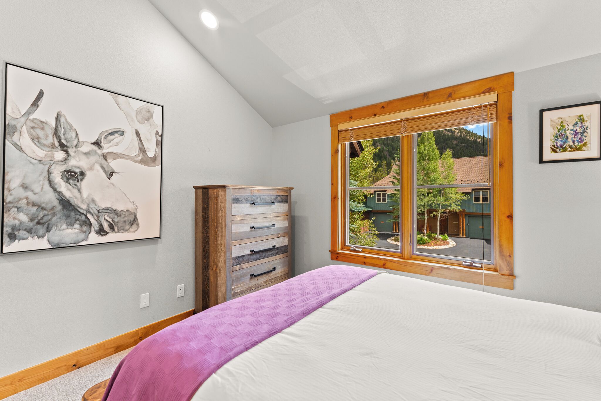 Een slaapkamer met een groot bed, een houten dressoir, een raam met uitzicht op een huis en een schilderij aan de muur.