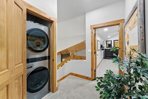A laundry room with two stacked washing machines, a wooden staircase, and a hallway leading to a room with a door and a plant. - St8709-3 (Keystone)
