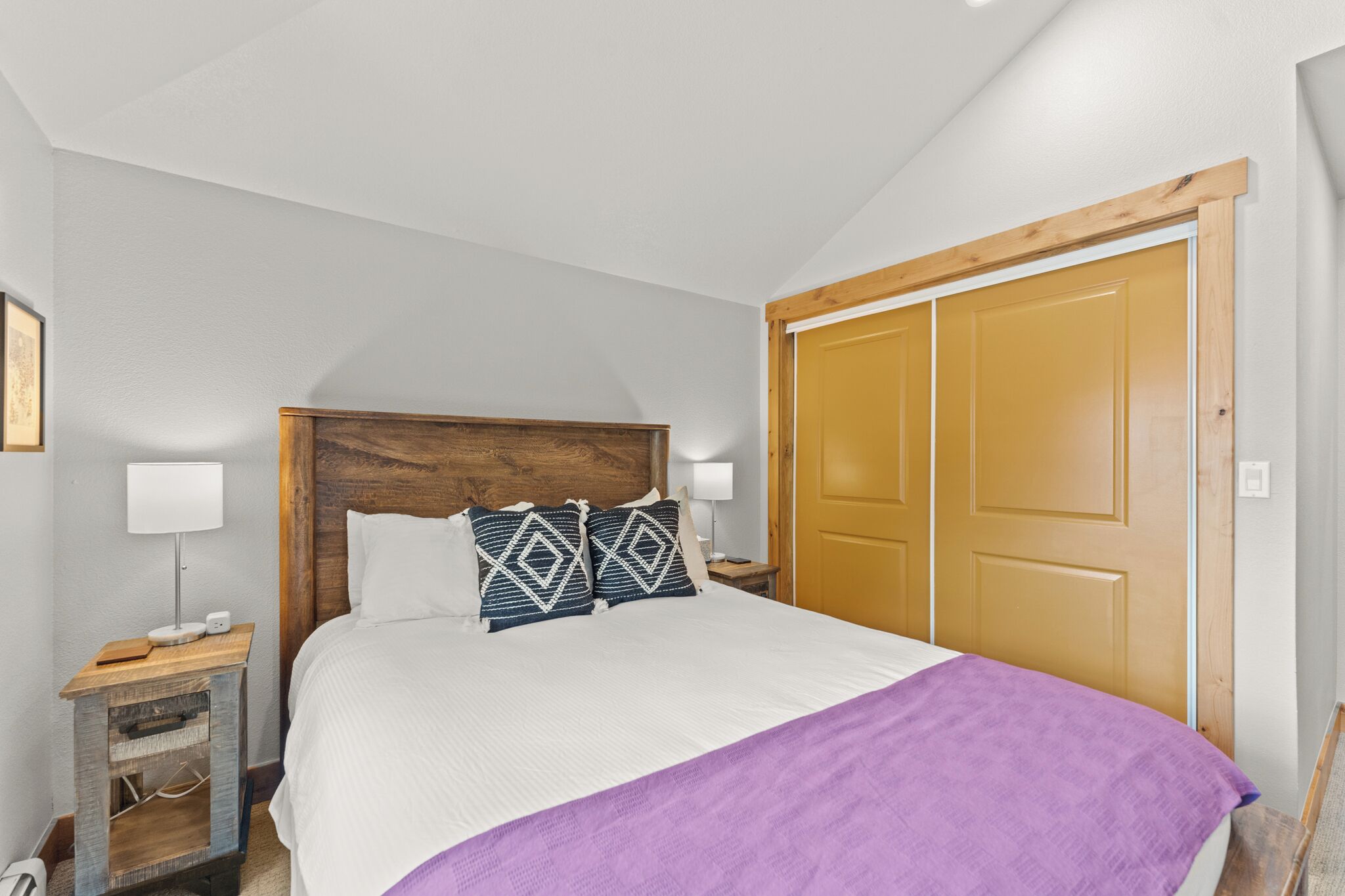 Un dormitorio con cabecera de madera, cama con ropa de cama blanca y morada, mesita de noche con lámpara y armario de madera.