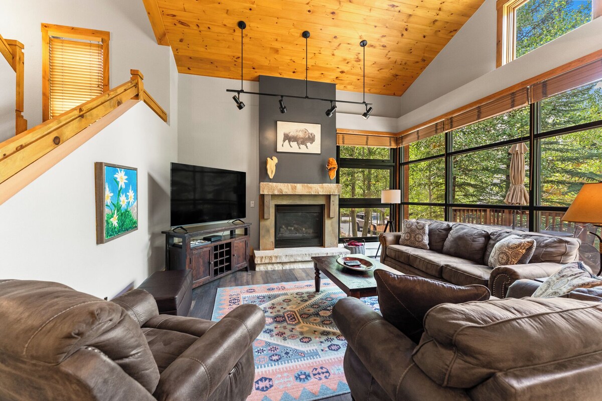 A cozy living room with a fireplace, leather sofas, and a large window offering a view of trees.