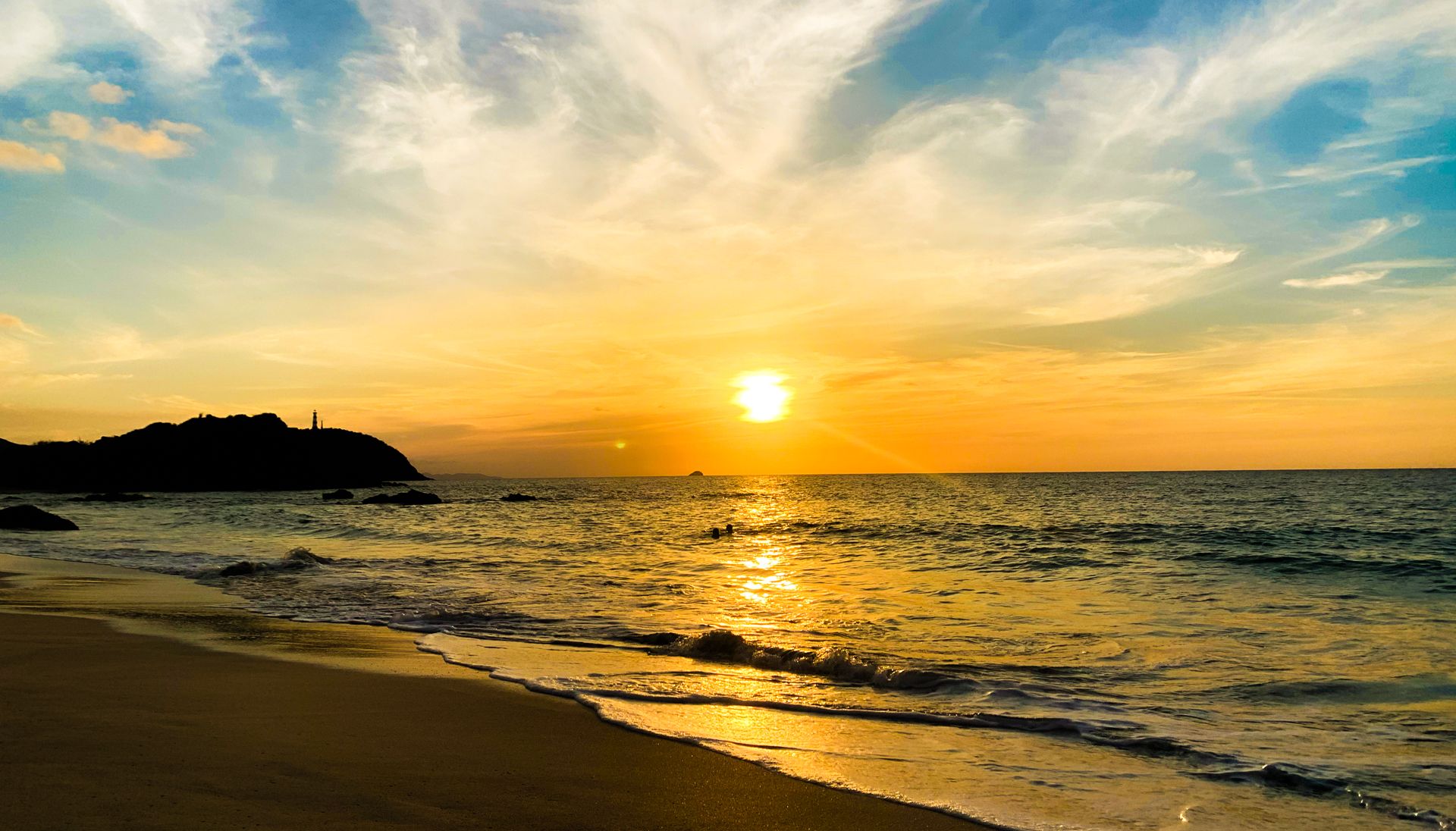 A sunset over a beach with waves, a distant island, and a lighthouse.