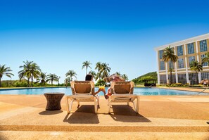 Zona junto a la piscina con sillones de descanso y un edificio al fondo.