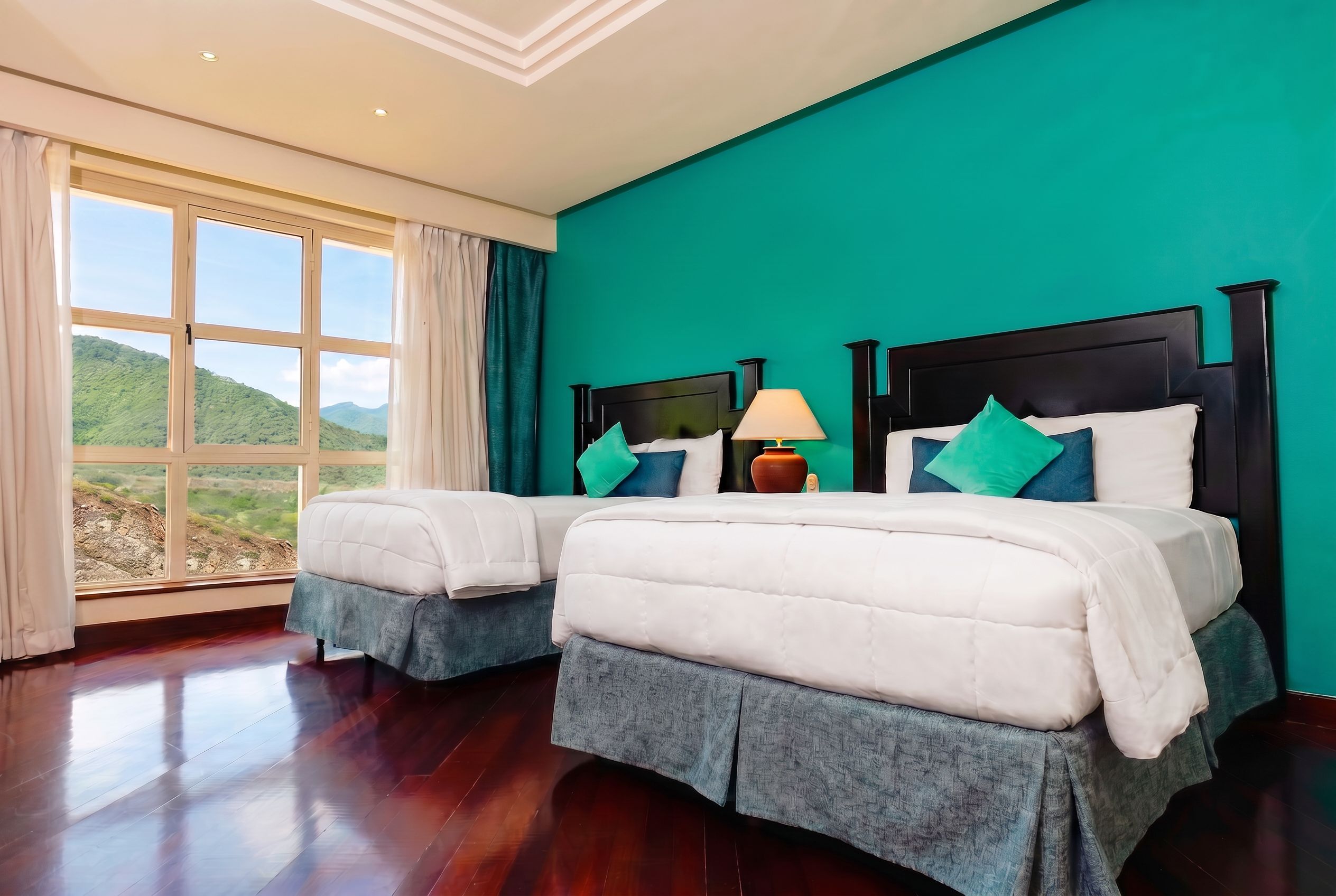 Habitación de hotel con dos camas, vista al exterior y una pared color turquesa.