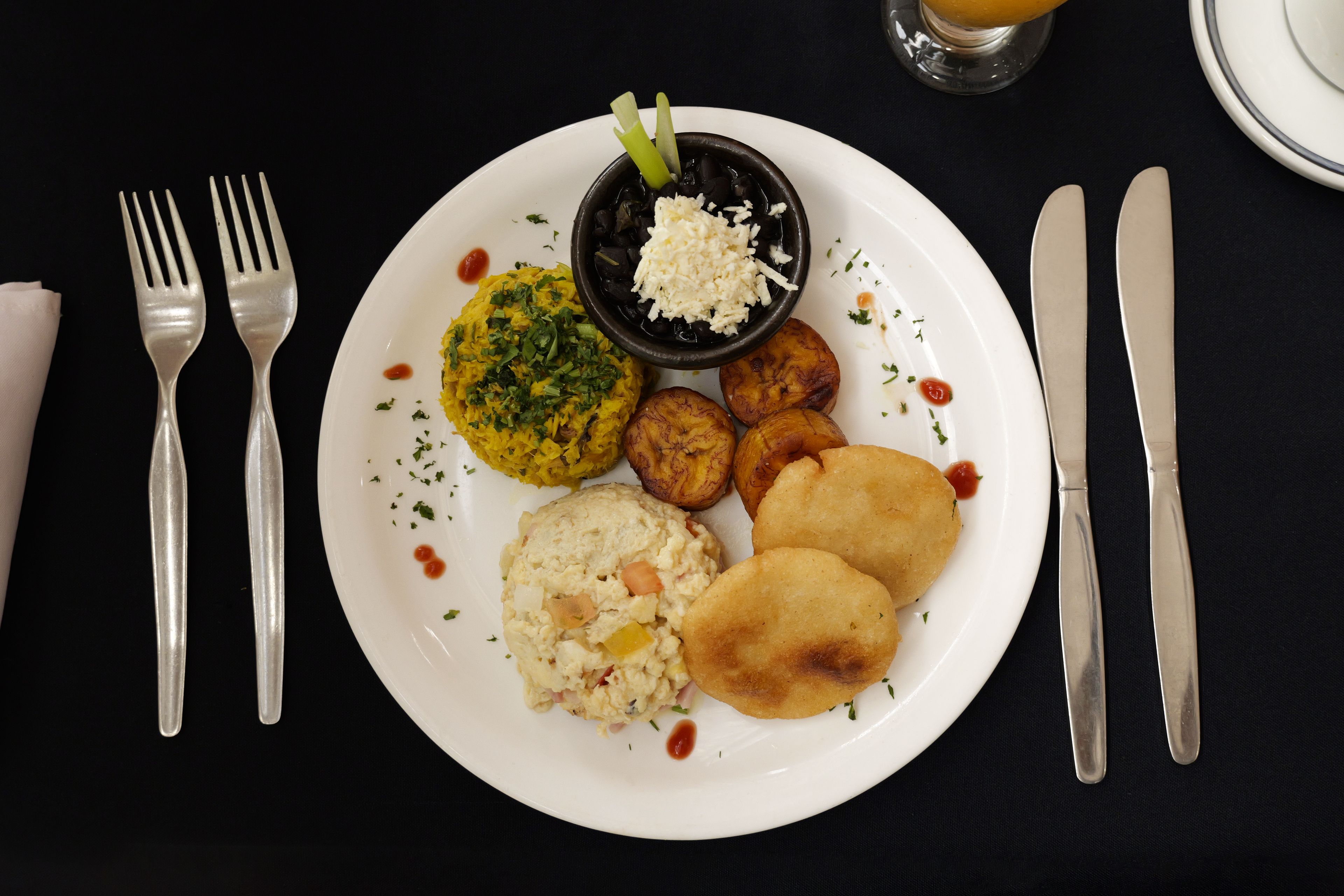 A plate of food with a variety of items including a yellow rice dish, a black bowl with a white topping, fried items, and a side of what appears to be a potato salad.