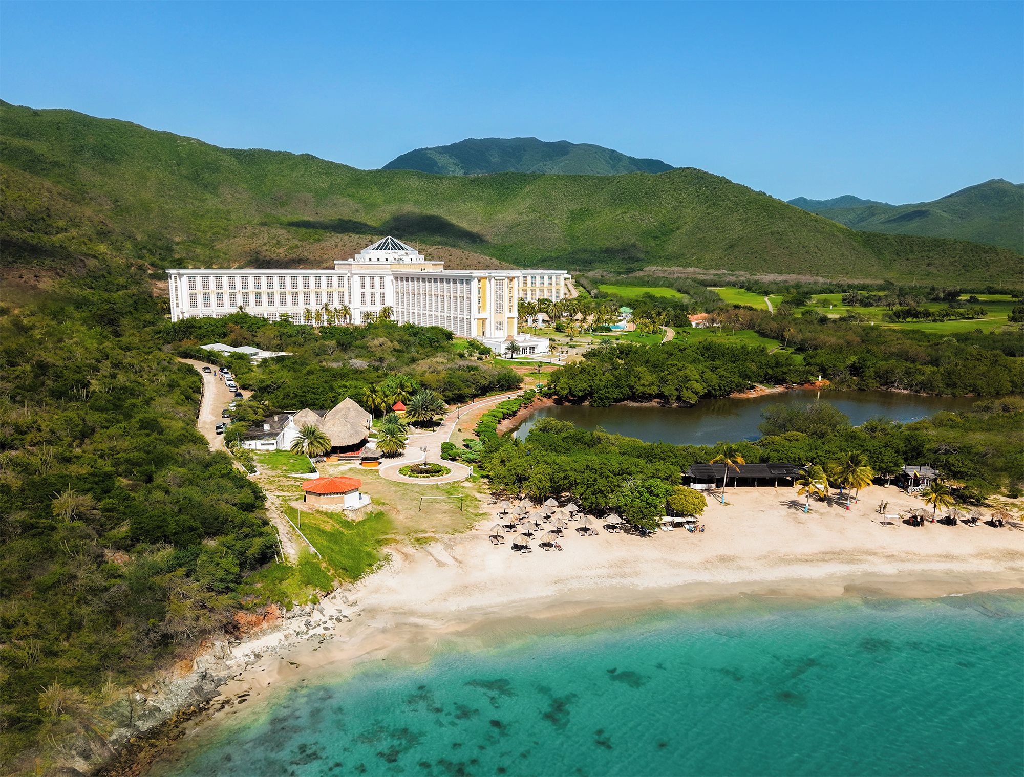 A large resort with a central building, a beach, and a mountainous backdrop.