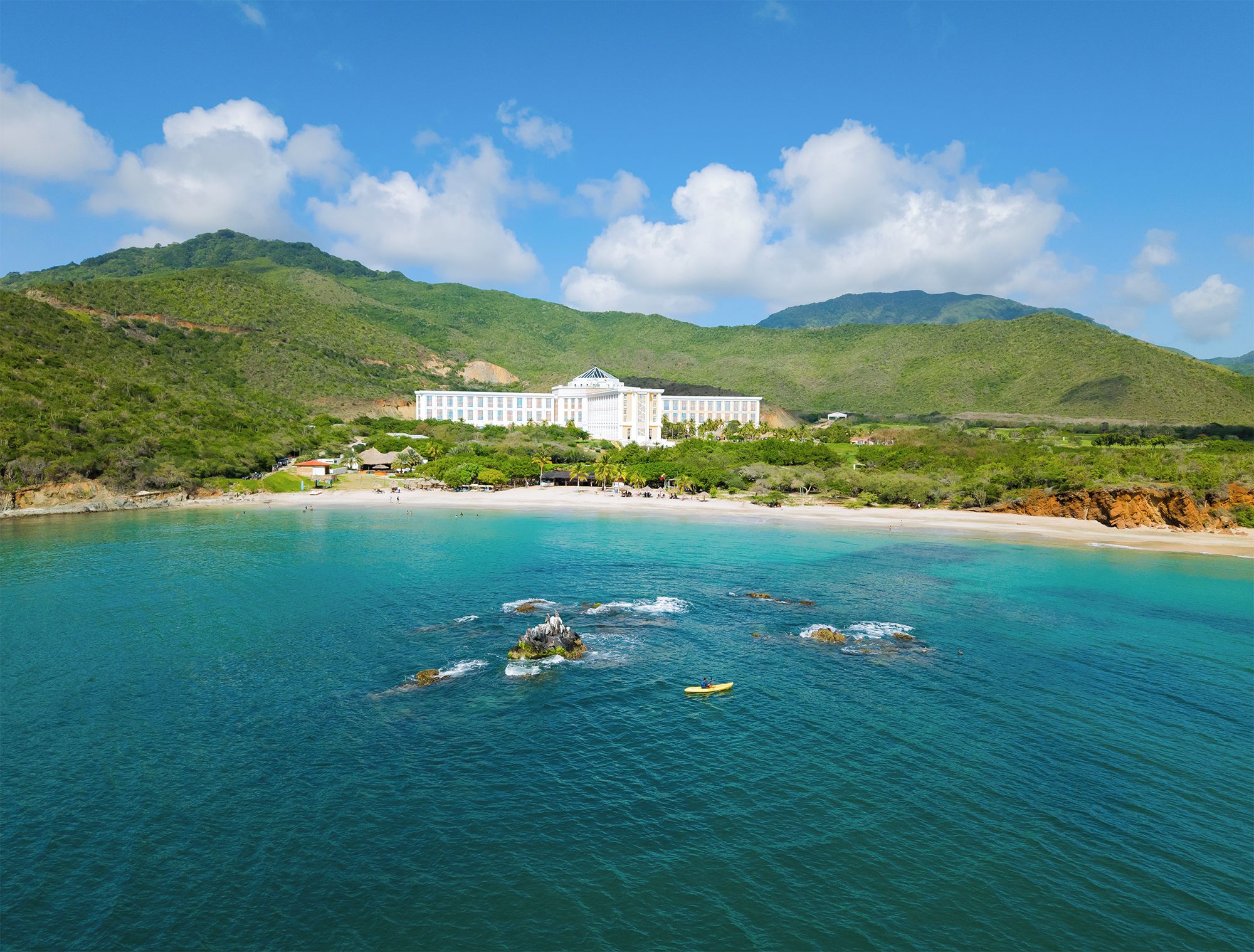Un hotel costero con un edificio grande, una playa y montañas al fondo.