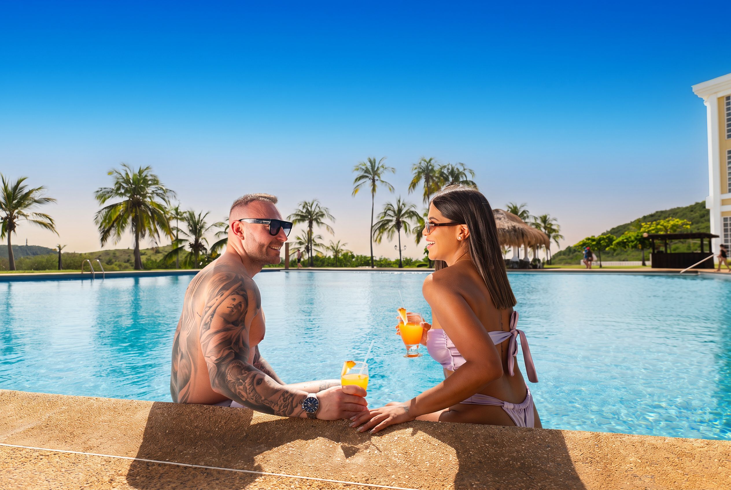 A couple by a pool with clear skies, palm trees, and a building in the background.
