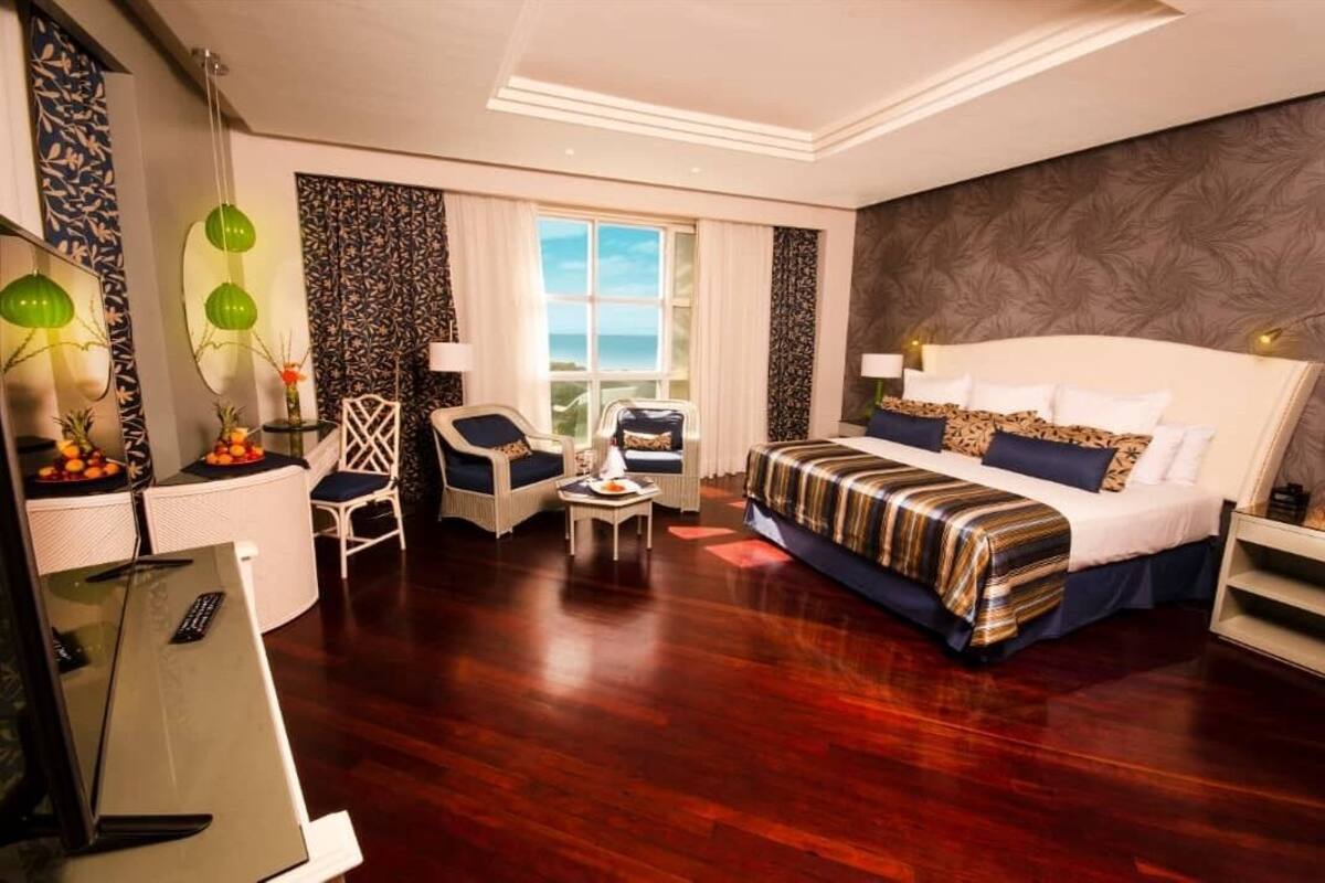 Ein Hotelzimmer mit einem großen Bett, einem Schreibtisch mit Computer, einem Sessel, einem kleinen Tisch mit Früchten und Meerblick durch das Fenster.