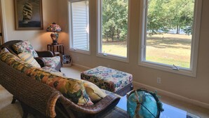 A living room with a wicker sofa, a matching ottoman, and a side table with a lamp. There are three large windows providing a view of a yard with trees. - Lakehouse on Ky Lake-Lakefront, Park like backyard, Covered Dock, Firepit. (Benton)