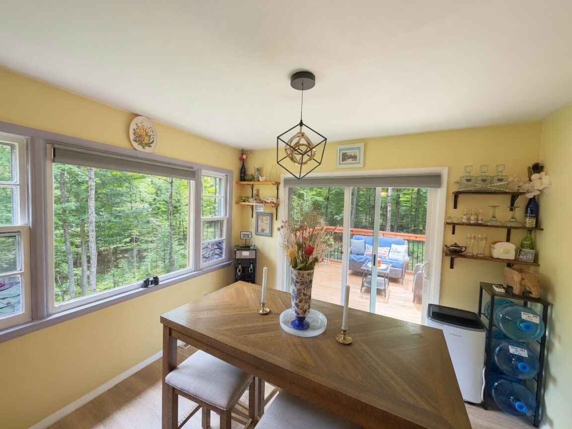 A dining area with a wooden table, a vase with flowers, and a view of the outdoors through large windows.