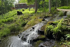 A forested area with a small waterfall, a wooden deck, and a building in the background. - Saratoga Excitement Meets Peaceful Retreat- 10 mins to the track! (Greenfield Center)