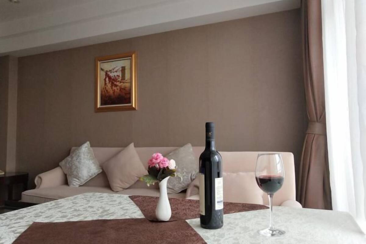 Una camera d'albergo con un divano, un vaso con fiori, una bottiglia di vino e un bicchiere da vino su un tavolo.