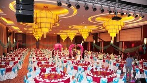 Ein Festsaal mit runden Tischen, bedeckt mit roten Tischdecken, und weißen Stühlen mit blauen Bändern, die in Reihen angeordnet sind.