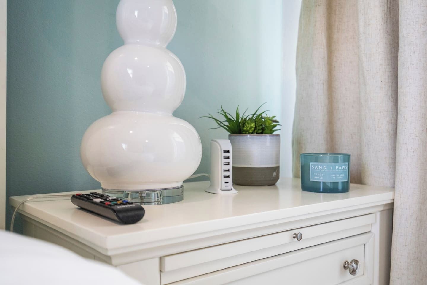A bedside table with a remote control, a white lamp, a potted plant, and a candle.