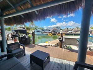 A covered deck with outdoor furniture, a pool, and boats in the background.