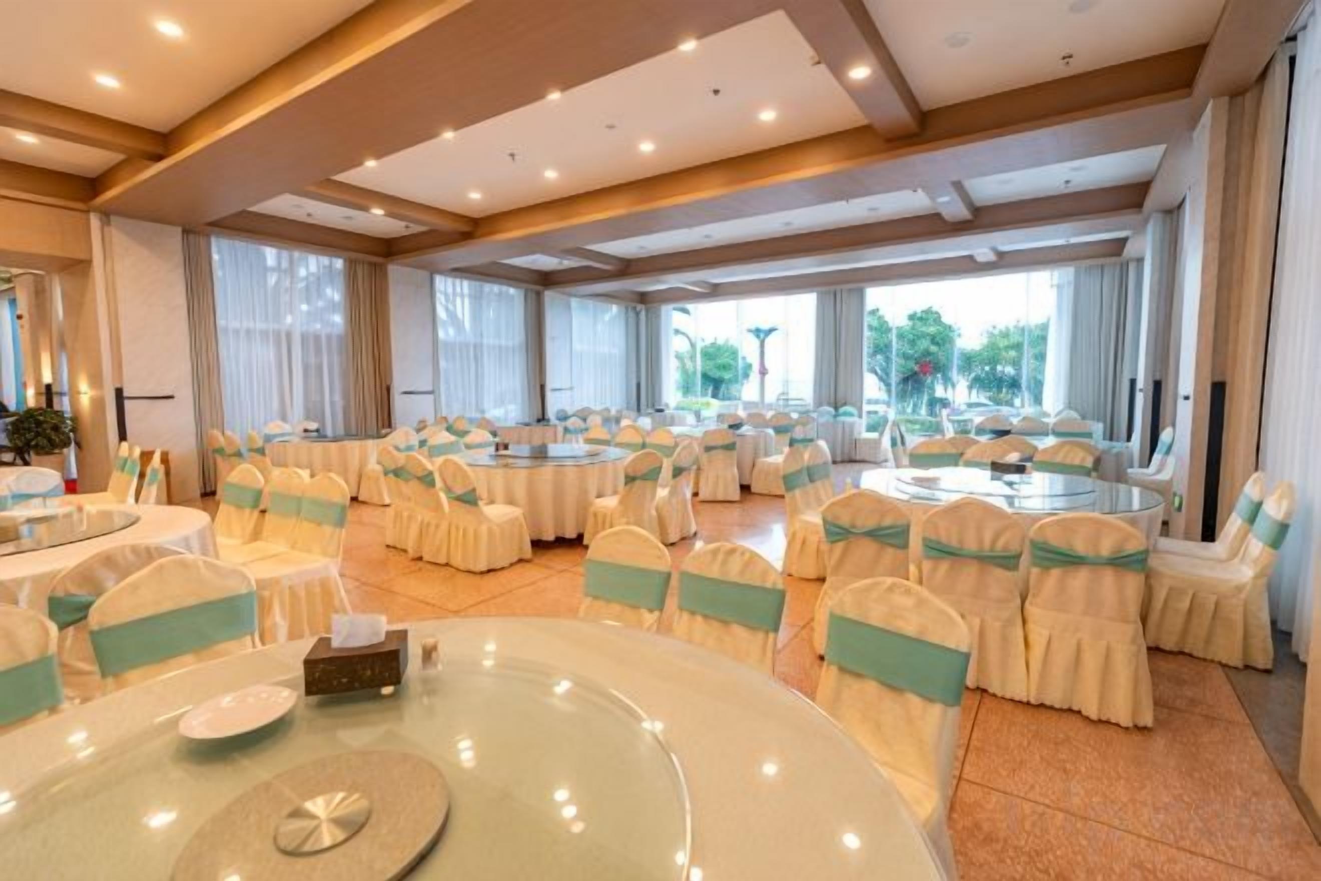 Un salón de conferencias con varias mesas redondas, cada una rodeada de sillas con cintas verdes, y grandes ventanales que ofrecen luz natural.