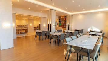 A modern dining area with multiple tables and chairs, a visible kitchen area, and a bookshelf.