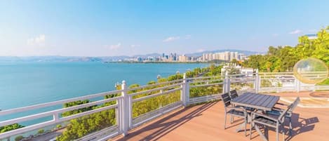 A wooden deck with a railing overlooking a body of water, a city skyline in the distance, and a clear sky.