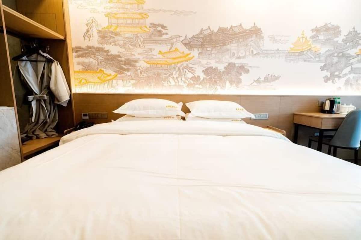A hotel room with a bed, pillows, a robe rack, a desk, and a wall with a traditional mural.