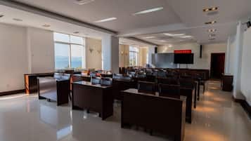 A conference room with rows of tables and chairs, a large screen at the front, and a view of the outdoors through large windows.