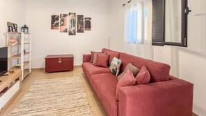 Una sala de estar con un sofá rojo, un aparador de madera, una estantería blanca con objetos decorativos y una alfombra estampada.