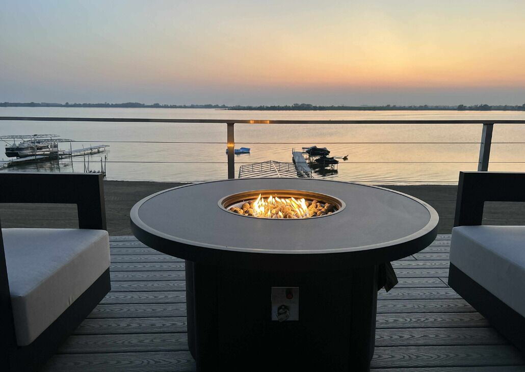 Une terrasse dotée d’un foyer circulaire, de deux canapés et offrant une vue sur une étendue d’eau paisible au coucher du soleil.