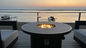A deck with a circular fire pit, two sofas, and a view of a calm body of water during sunset.