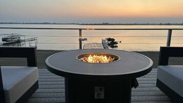 Une terrasse dotée d’un foyer circulaire, de deux canapés et offrant une vue sur une étendue d’eau paisible au coucher du soleil.