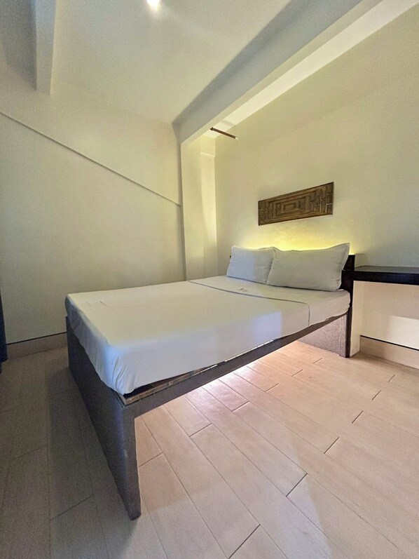 A single bed with a wooden frame and white bedding in a room with light wooden flooring and white walls. - Selah Lofts Pasay (Pasay)