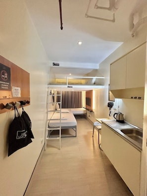 A compact hotel room with bunk beds, a kitchenette, a small table, and a wall-mounted shelf with bags. - Selah Lofts Pasay (Pasay)