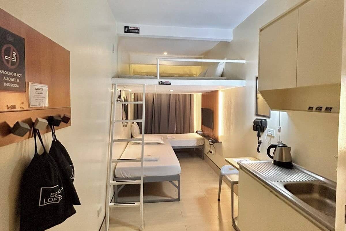A compact hotel room with bunk beds, a kitchenette, a small table, and a wall-mounted shelf with bags.