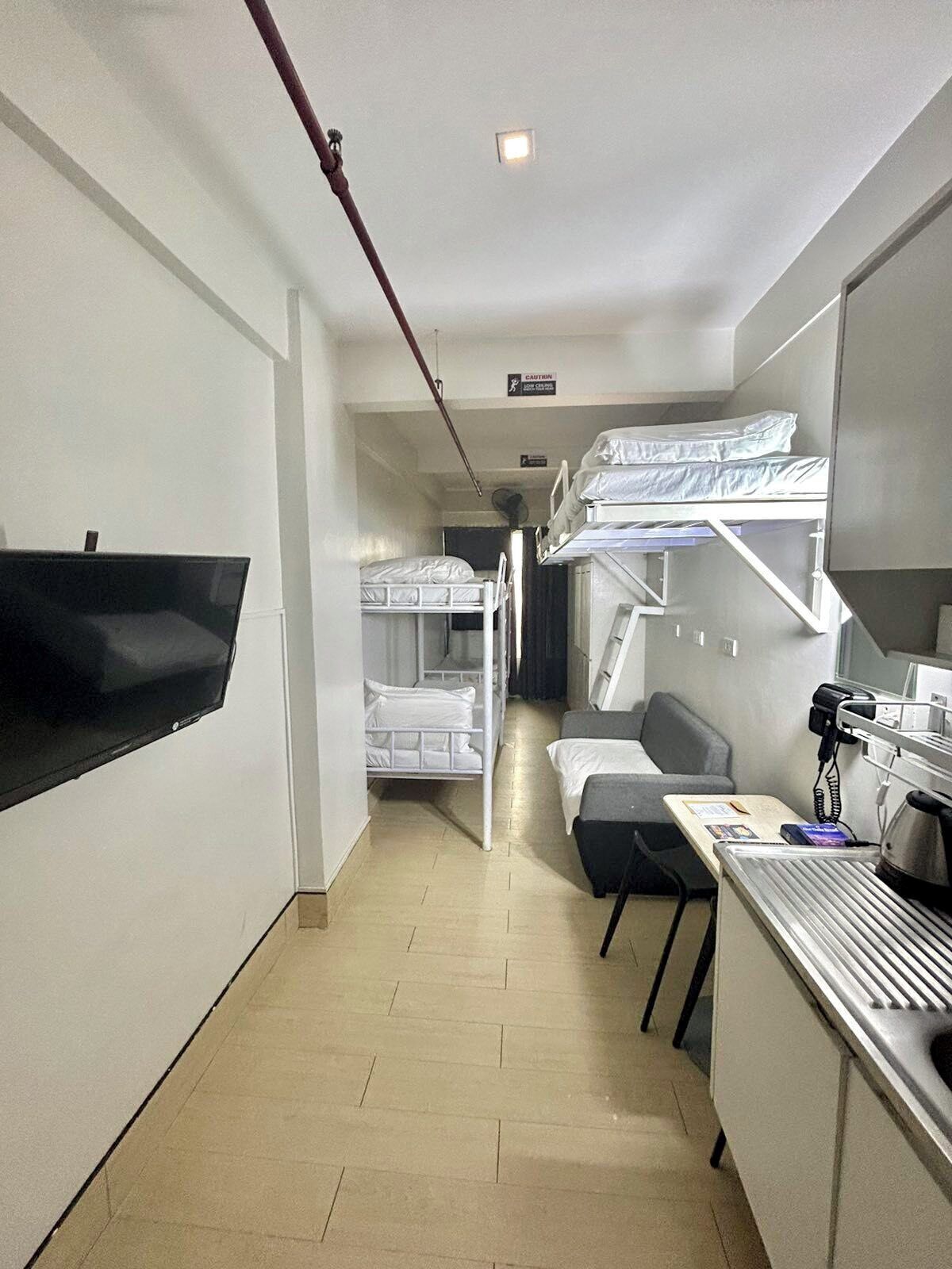 Une chambre d’hôtel avec des lits superposés, un bureau avec un téléphone, un téléviseur à écran plat fixé au mur et une petite kitchenette.
