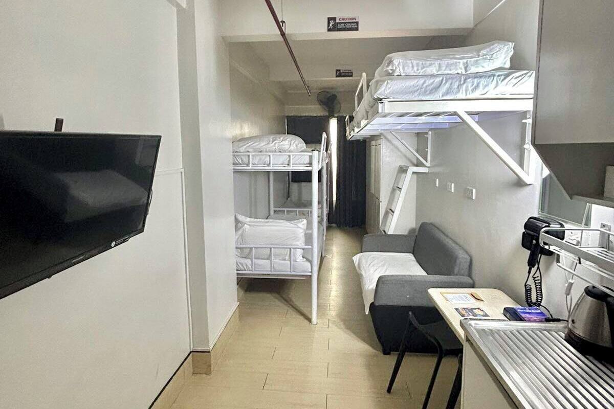 A hotel room with bunk beds, a desk with a telephone, a flat-screen TV mounted on the wall, and a small kitchenette.