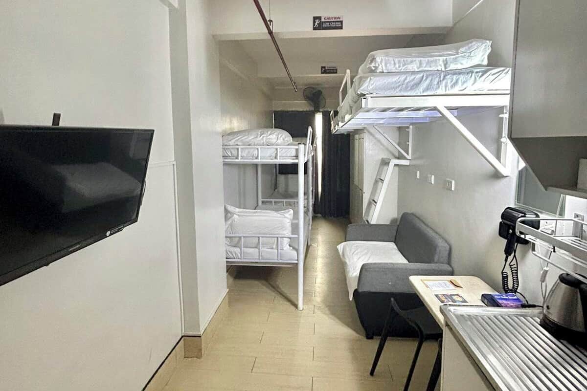 A hotel room with bunk beds, a desk with a telephone, a flat-screen TV mounted on the wall, and a small kitchenette.