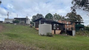 A two-story house with a stone exterior, a large water tank, and a green-roofed structure on the right. - Currawong Cottage - Fishing Paradise (Anglers Reach)