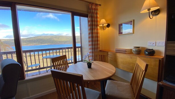 Una habitación con una mesa de comedor de madera, sillas, una orquídea en maceta, vista al mar y un balcón con barandilla.