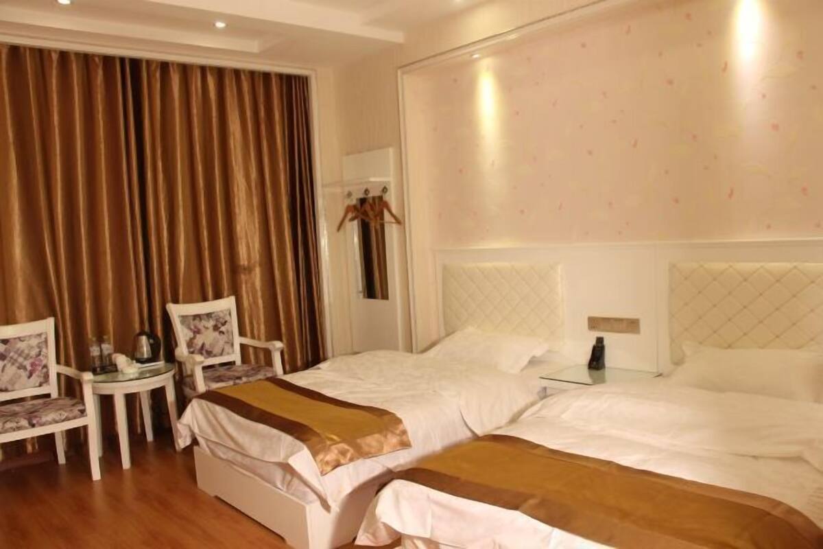 A hotel room with two beds, a small table, two chairs, a wardrobe, and a large window with curtains.