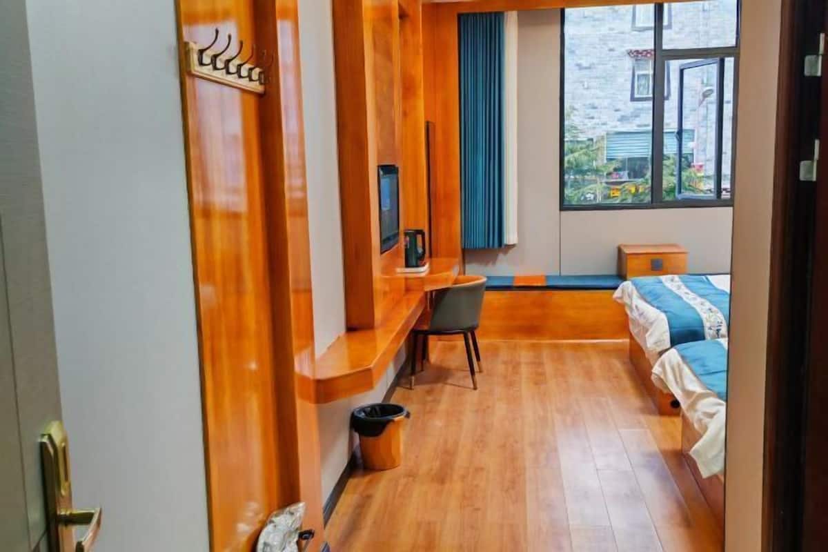 A hotel room with a wooden desk, a bed with blue bedding, a window with a view of the outside, and a small wooden nightstand.