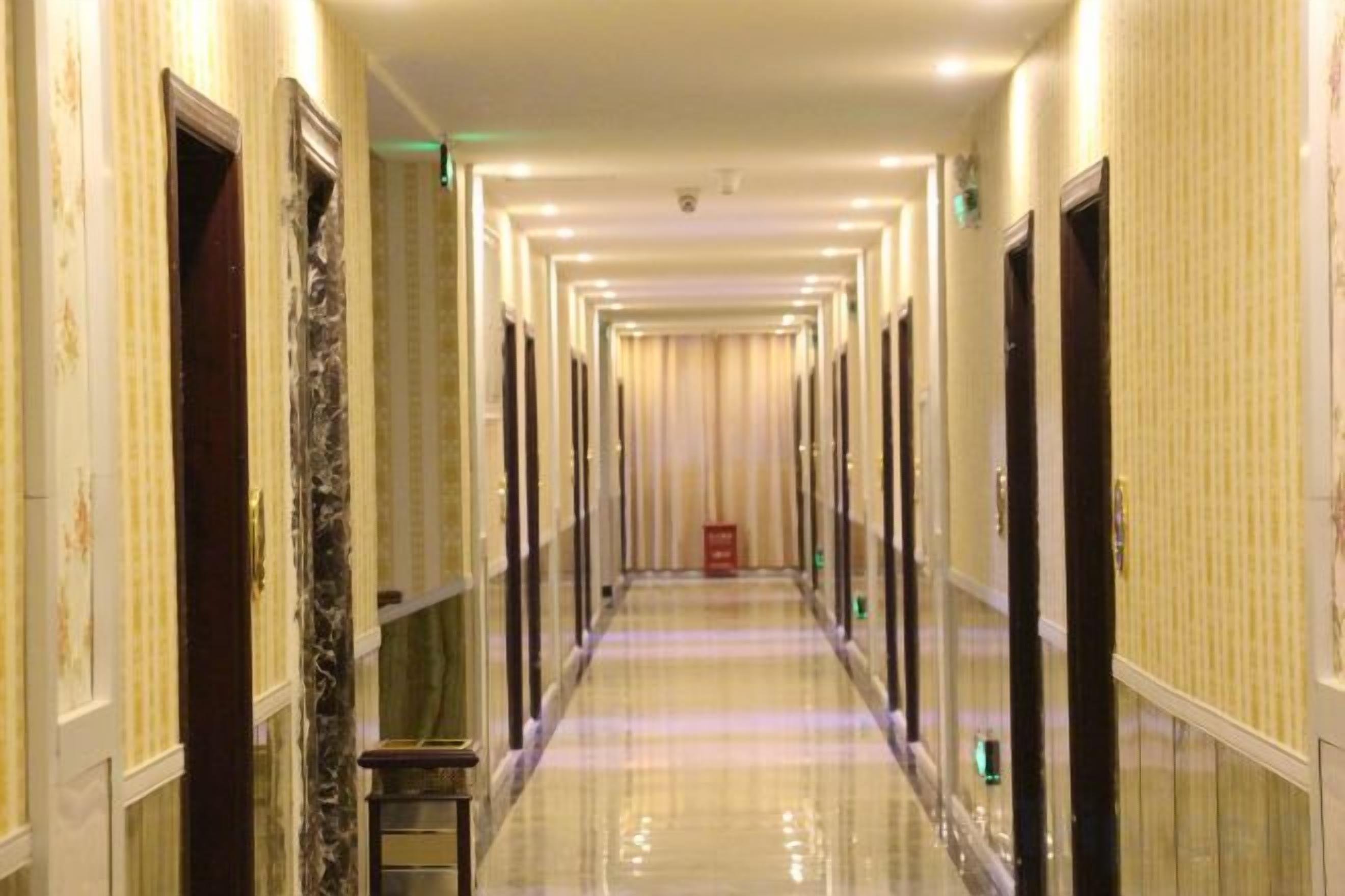 Corredor comprido e estreito de um hotel, com portas em ambos os lados, chão polido e um objeto vermelho à direita.