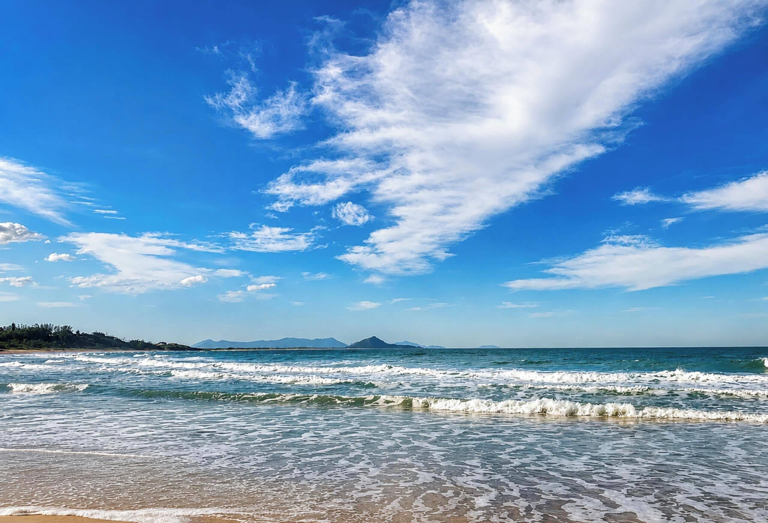 Uma praia com ondas, céu claro e montanhas ao longe.