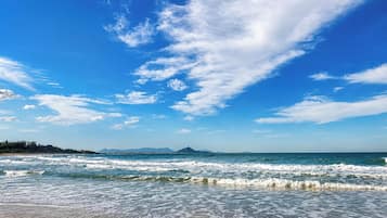 Uma praia com ondas, céu claro e montanhas ao longe.