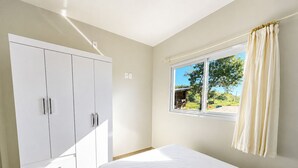 A bedroom with a bed, a wardrobe, a window with curtains, and a view of greenery outside. - Paraíso do Coral - Vista Mar - Garopaba (Paulo Lopes)