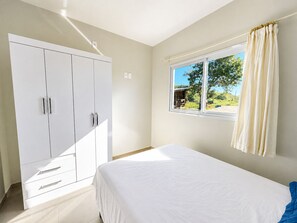 Ein Schlafzimmer mit Bett, Kleiderschrank, einem Fenster mit Vorhängen und Blick auf Grünflächen.