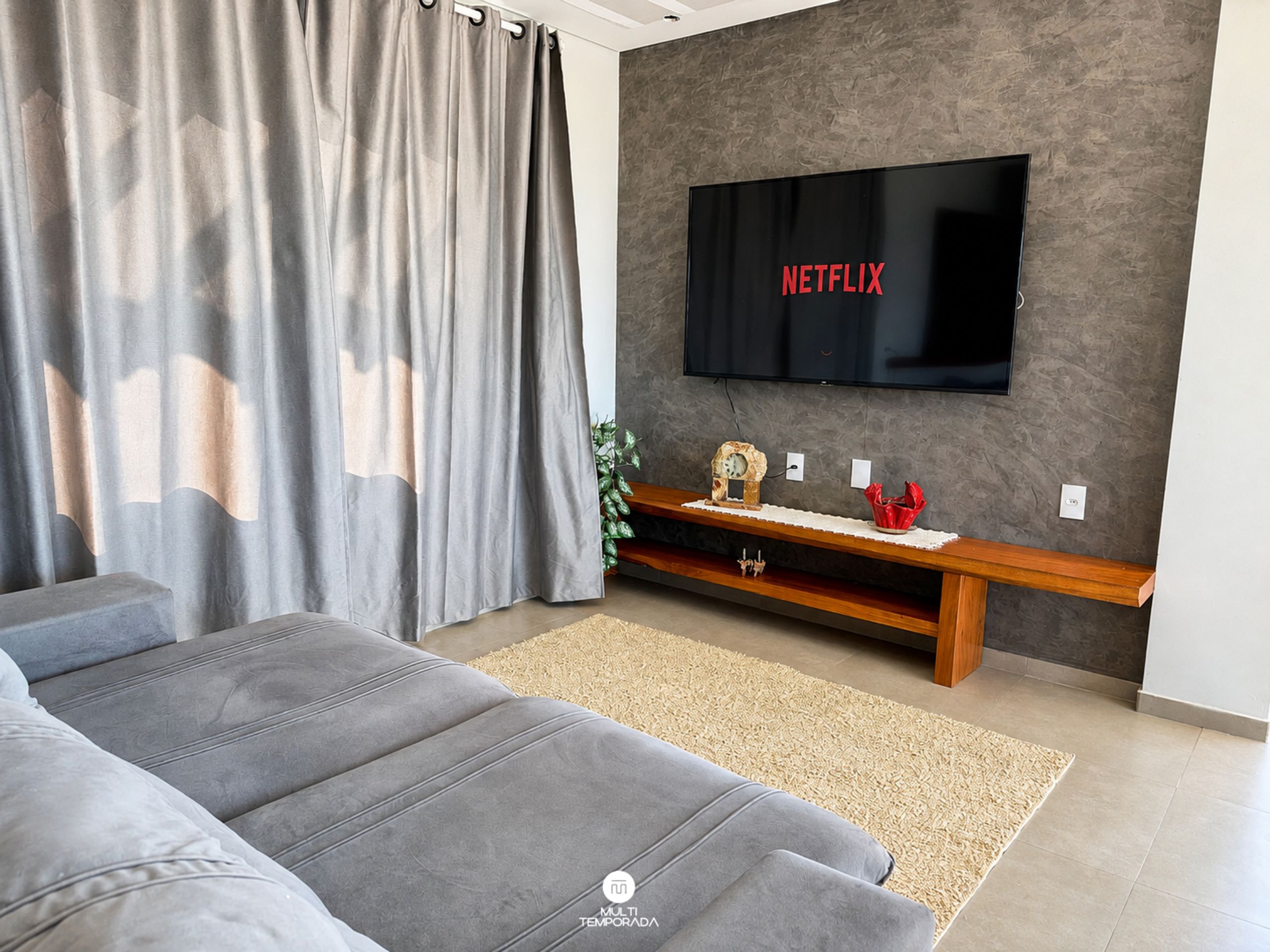 A modern living room with a grey sofa, a flat-screen TV mounted on a textured wall, and a wooden entertainment unit.