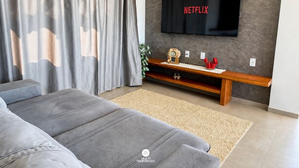 A modern living room with a grey sofa, a flat-screen TV mounted on a textured wall, and a wooden entertainment unit. - Paraíso do Coral - Vista Mar - Garopaba (Paulo Lopes)