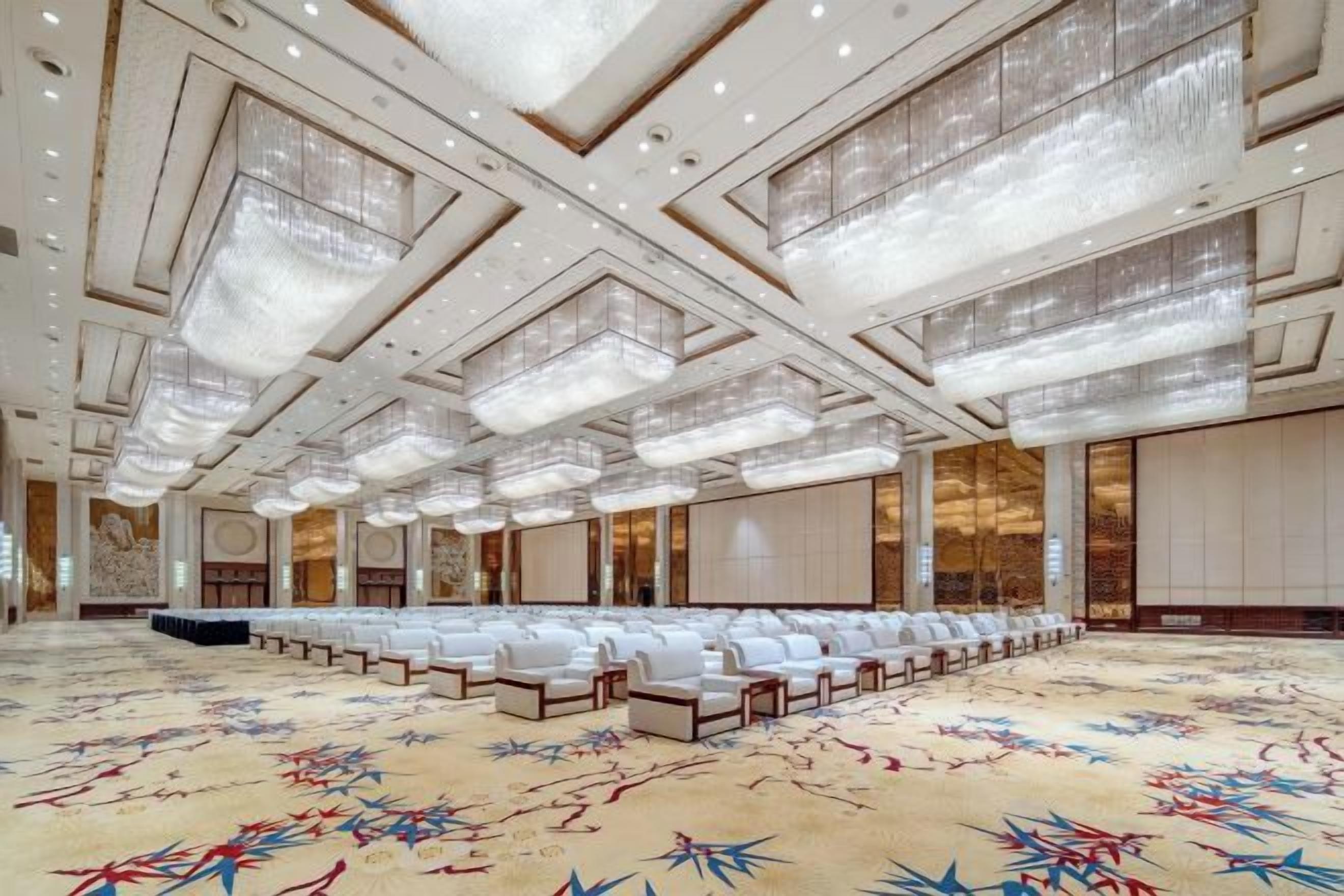 A spacious conference room with rows of white chairs, a patterned carpet, and a high ceiling with chandeliers.